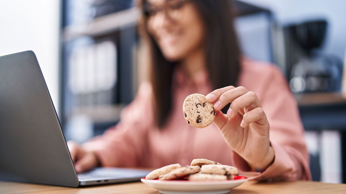Sweets sales: A young woman sitting in front of a laptop laughing, while taking a biscuit from a pile of biscuits on a plate.