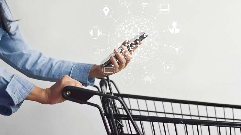 Close-up of a person holding a smartphone in one hand and pushing a shopping cart with the other